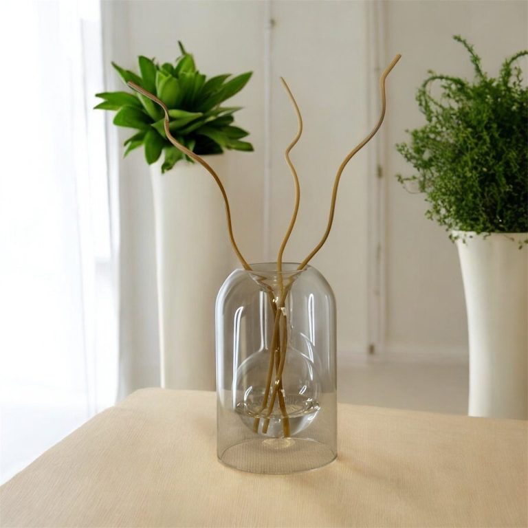 Vase transparent avec des tiges dorées et une plante verte, sur une table claire.